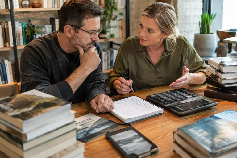 Author reviewing a budget sheet beside books and a laptop